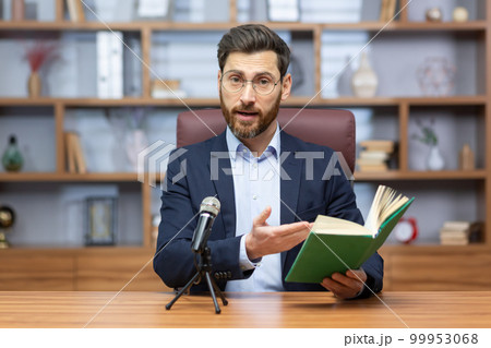 Senior mature pastor reading bible book online, man looking at camera sitting in home office and recording audio book online. Senior mature pastor reading bible book online, man looking at camera sitting in home office and recording audio book online. 99953068