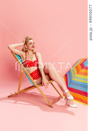 Summer vacation. Portrait of beautiful young woman in swimming suit posing against pink studio background with sun umbrella. Concept of beauty and pin-up style 99956077