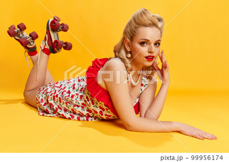 Leisure time. Portrait of beautiful young girl with stylish hairstyle in dress, on rollers posing against yellow studio background. Concept of beauty and pin-up style 99956174