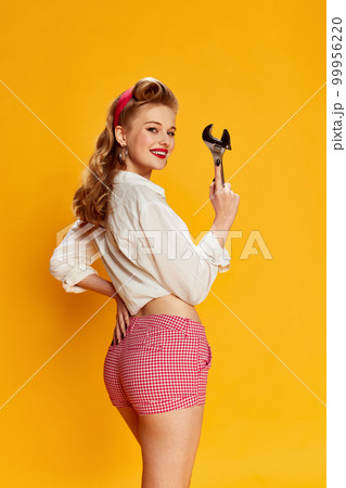 Portrait of beautiful young blonde girl in white shirt and pink shorts posing with tool, wrench against yellow studio background. Concept of beauty and pin-up style Portrait of beautiful young blonde girl in white shirt and pink shorts posing with tool, wrench against yellow studio background. Concept of beauty and pin-up style 99956220