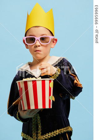 Serious little cute boy in costume of medieval little prince in paper crown tasting popcorn over light blue background. Concept of children emotions, eras comparison, vintage fashion Serious little cute boy in costume of medieval little prince in paper crown tasting popcorn over light blue background. Concept of children emotions, eras comparison, vintage fashion 99959963