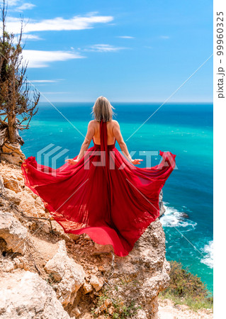 A girl with loose hair in a long red dress descends the stairs between the yellow rocks overlooking the sea. A rock can be seen in the sea. Sunny path on the sea from the rising sun 99960325
