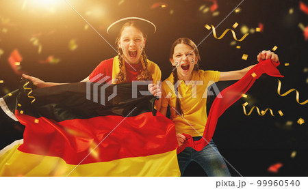 Young girls emotionally watching football match, holding flag and cheering team over dark background with confetti 99960540