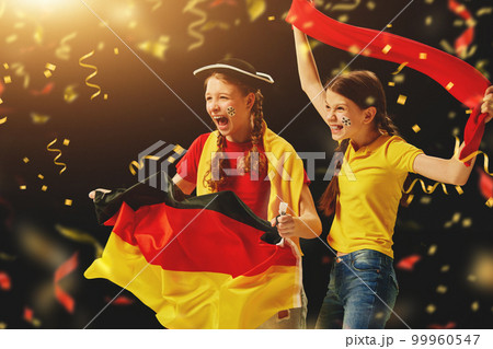 Excited young girls, teens emotionally cheering up favourite german football team on championship, game over dark background with confetti Excited young girls, teens emotionally cheering up favourite german football team on championship, game over dark background with confetti 99960547