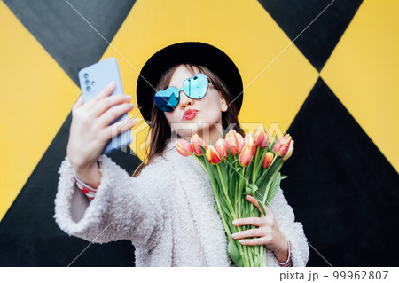 Emotional young fashion woman in heartshaped sunglasses holding bouquet of tulip flower, taking selfie photo on geometry bright yellow and black wall background. Urban city street fashion. Spring mood Emotional young fashion woman in heartshaped sunglasses holding bouquet of tulip flower, taking selfie photo on geometry bright yellow and black wall background. Urban city street fashion. Spring mood 99962807