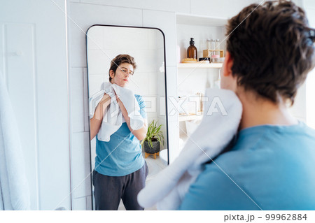 Concentrated caucasian teenage boy with acne problem takes care of his face skin at home. He looking in mirror and wipes face with towel in bathroom. Teenager skin care every day treatment process. 99962884