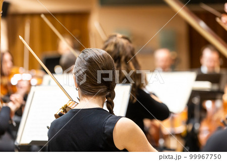 Professional symphonic string orchestra performing on stage and playing a classical music concert, violinist in a foreground Professional symphonic string orchestra performing on stage and playing a classical music concert, violinist in a foreground 99967750