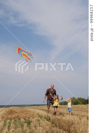 A father and his daughters launch a rainbow kite into a clear sky. Kids have fun with Dad in the field on a summer day. A father and his daughters launch a rainbow kite into a clear sky. Kids have fun with Dad in the field on a summer day. 99968257