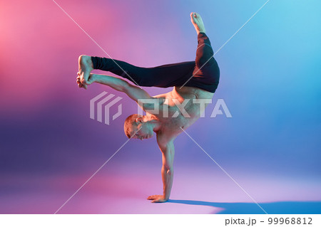 Young handsome acrobat or gymnast training to keep his body balance, standing on one hand while being barefoot and topless, touching his leg, showing his power. 99968812