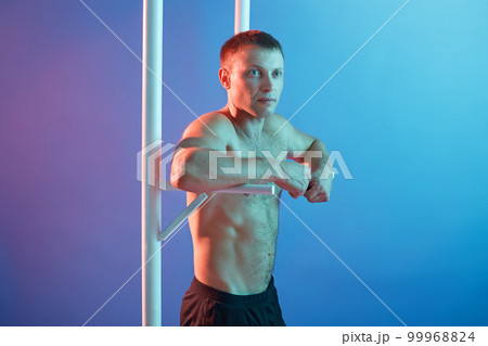 Handsome young man doing exercise on horizontal bar, guy posing topless, keeps fit, posing isolated over neon background, male looking aside with calm expression. 99968824