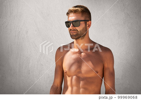 Waist up portrait of attractive macho man with bristle in trendy eyewear, standing naked against grey blank wall, looking aside with confident expression, demonstrating his suntanned, sport body Waist up portrait of attractive macho man with bristle in trendy eyewear, standing naked against grey blank wall, looking aside with confident expression, demonstrating his suntanned, sport body 99969068
