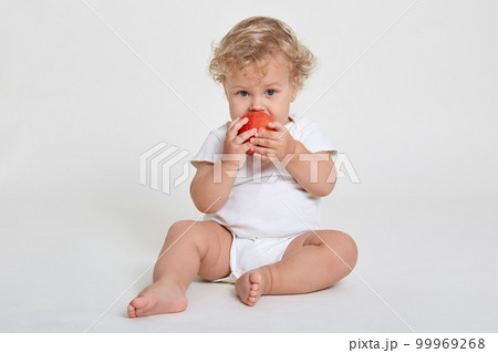 Beautiful blond curly haired little boy eats fresh red apple, healthy breakfast, starts day with healthy food, infant wearing body suit sitting barefoot on floor against white wall. Beautiful blond curly haired little boy eats fresh red apple, healthy breakfast, starts day with healthy food, infant wearing body suit sitting barefoot on floor against white wall. 99969268