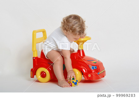 Cute kid boy on light background sitting barefoot on tolocar machine, lifting his toy car off floor, wearing body suit, has blond wavy hair, playing with his toys isolated over white background. 99969283