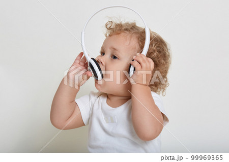 Charming baby boy with blond hair playing with headphones, biting it, wearing white t shirt, looking away, curious kid having fun, listening music. Charming baby boy with blond hair playing with headphones, biting it, wearing white t shirt, looking away, curious kid having fun, listening music. 99969365