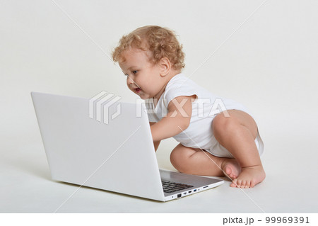 Adorable baby sitting on floor and looking at laptop screen with exited facial expression, infant wearing white body suit, having blond curly hair , touching keyboard with hand. 99969391