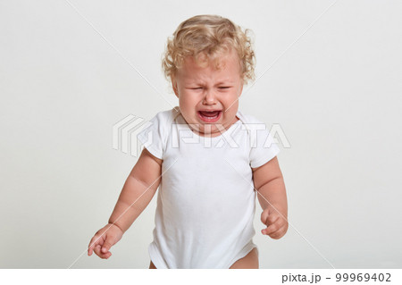 Portrait of sad upset little boy crying, looking down, frowning face, wearing bodysuit, being in bed mood, feels hungry, posing isolated over white background. 99969402