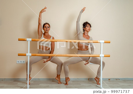 Near the barre, in the choreographic hall, two women in a beige top and leggings are engaged. Near the barre, in the choreographic hall, two women in a beige top and leggings are engaged. 99971737