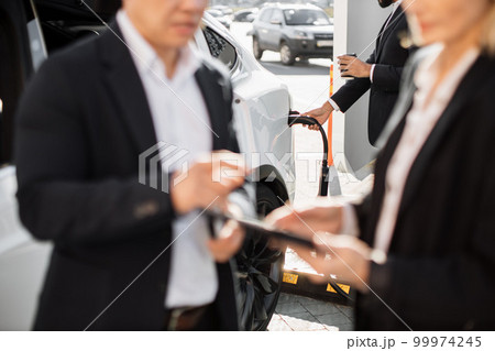 Business people using gadgets and charging electric car Business people using gadgets and charging electric car 99974245