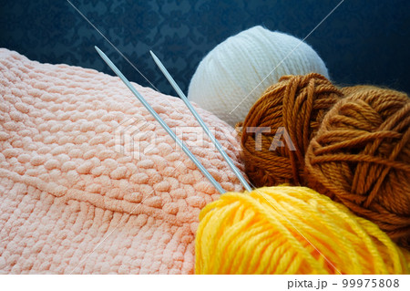 Two knitting needles stick out from a ball of yarn in the background of a piece of knitted goods. Yellow, coral pink, brown, white yarns of different thicknesses and compositions. Close-up, copy space Two knitting needles stick out from a ball of yarn in the background of a piece of knitted goods. Yellow, coral pink, brown, white yarns of different thicknesses and compositions. Close-up, copy space 99975808
