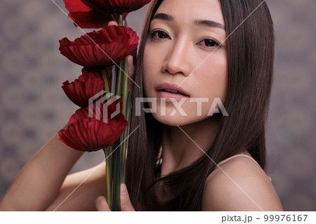 Cosmetology model with perfect skin posing with poppies bouquet in hands, looking at camera with serious facial expression. Beautiful asian woman standing with red flowers for cosmetics brand Cosmetology model with perfect skin posing with poppies bouquet in hands, looking at camera with serious facial expression. Beautiful asian woman standing with red flowers for cosmetics brand 99976167