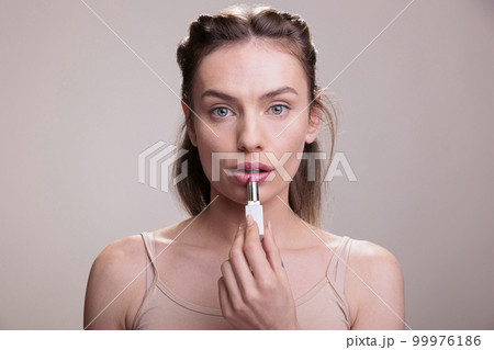 Young woman applying pink lipstick and doing nude make up portrait. Natural beautiful cosmetic product model using color lipbalm tube, showing lips makeup routine and looking at camera 99976186