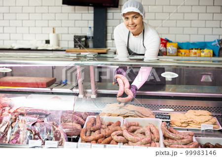 Happy female butcher holding large beef sausage in meat section of supermarket Happy female butcher holding large beef sausage in meat section of supermarket 99983146