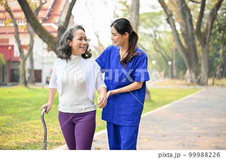 Lovely female caregiver assisting and supporting an old lady to walk with a walking stick 99988226