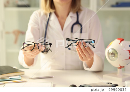 Ophthalmologist holds different eyeglasses in her hands while sitting at desk in medical clinic. Ophthalmologist holds different eyeglasses in her hands while sitting at desk in medical clinic. 99990035