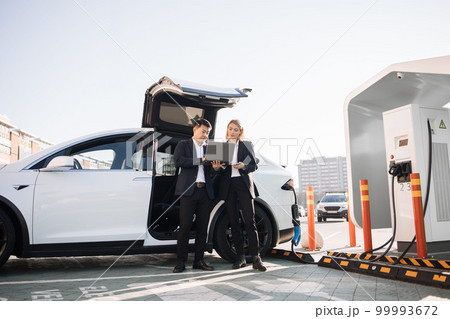 Business colleagues using laptop while charging electric car Business colleagues using laptop while charging electric car 99993672