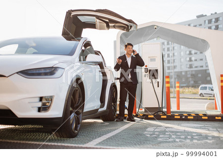 Asian man talking on mobile while charging electric car 99994001