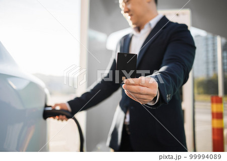Asian man holding credit card and charging car outdoors Asian man holding credit card and charging car outdoors 99994089