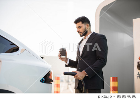 Businessman with coffee in hand charging modern car 99994696