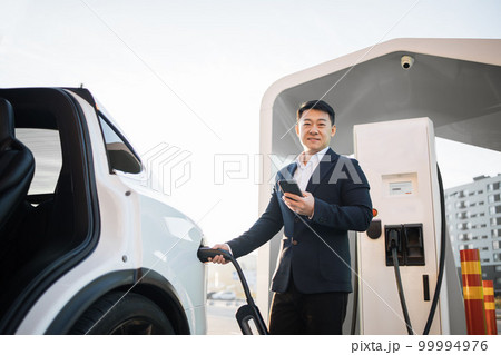 Asian man charging electro car and using smartphone Asian man charging electro car and using smartphone 99994976