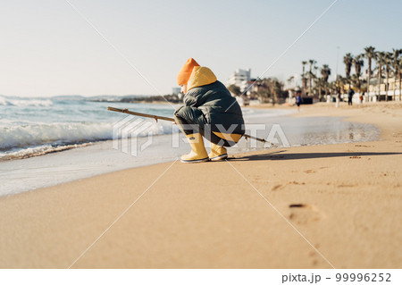 Boy in yellow rubber boots playing with stick and sand at the beach. School kid touching water at autumn winter sea. Child having fun with waves at the shore. Spring Holiday vacation concept 99996252