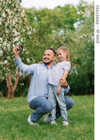 Dad and his six-year-old daughter take selfies on the phone, smiling and having fun on camera in the park in summer or spring Dad and his six-year-old daughter take selfies on the phone, smiling and having fun on camera in the park in summer or spring 99997092