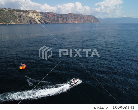 Happy people swim on air mattress behind a high speed water bike. Tourists ride on inflatable watercraft boat. Sea attraction. Water scooter skating people on large Inflatable raft. Aerial copter view 100003341