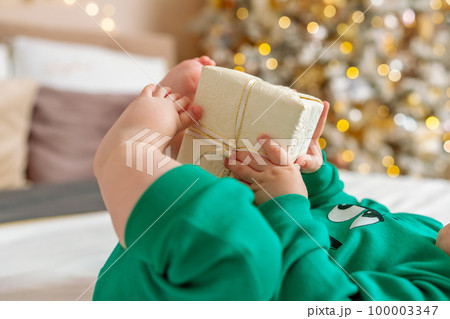 Baby gift. A little boy in a green suit holds a gift box with his feet and hands in bed. Baby gift. A little boy in a green suit holds a gift box with his feet and hands in bed. 100003347