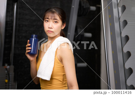 ドリンクを持ちながら視線を向けるスポーツウェア姿の若い女性 100011416