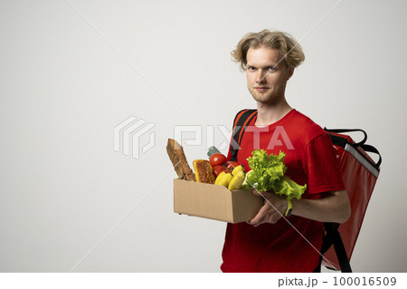 Happy delivery man in red uniform with a box full of groceries over white background. Express delivery, food delivery, online shopping concept. Happy delivery man in red uniform with a box full of groceries over white background. Express delivery, food delivery, online shopping concept. 100016509