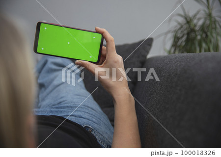 Back view of young europen woman who is using chroma key smartphone screen.She is sitting on the sofa in her cozy living room. Back view of young europen woman who is using chroma key smartphone screen.She is sitting on the sofa in her cozy living room. 100018326