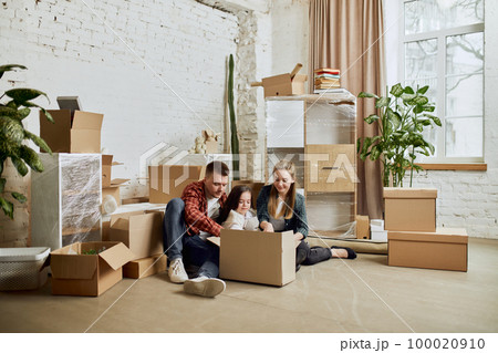 Young happy family, man, woman and little girl sitting in living room on daytime. Moving into new flat, apartment with many cardboard boxes Young happy family, man, woman and little girl sitting in living room on daytime. Moving into new flat, apartment with many cardboard boxes 100020910