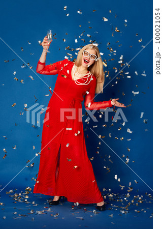 Portrait of beautiful woman in stylish red dress posing with champagne against blue studio background with confetti. Party and celebration 100021054