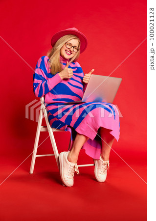 Happy smiling woman in colorful stylish clothes sitting on chair and having online video call on laptop against red studio background Happy smiling woman in colorful stylish clothes sitting on chair and having online video call on laptop against red studio background 100021128