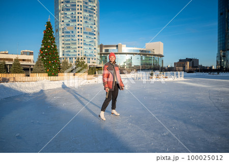 a young beautiful girl on skates in a sheepskin coat on a skating rink against the backdrop of the metropolis and the rays of the sun 100025012