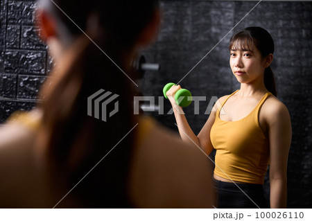 鏡を見ながらダンベルを使ってトレーニングをする若い女性 100026110
