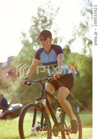 Smiling middle aged woman in cap with glasses cycling in summer park, cycling cardio training, city walks with bike, weight control 100033021