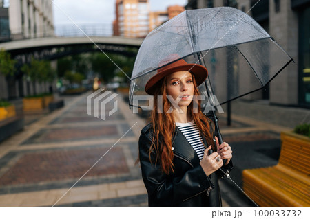 Portrait of elegant young woman in fashion hat standing on beautiful city street with transparent umbrella enjoying rainy weather outdoors, smiling looking at camera. Concept of female lifestyle. 100033732
