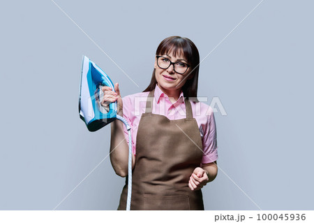 Woman in apron posing with iron, on gray background 100045936