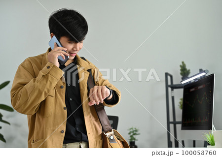 Image of young businessman or trader checking time on his watch and talking on mobile phone in modern office Image of young businessman or trader checking time on his watch and talking on mobile phone in modern office 100058760