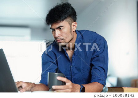 Portrait of an Asian man working on a computer and drinking coffee. 100059142